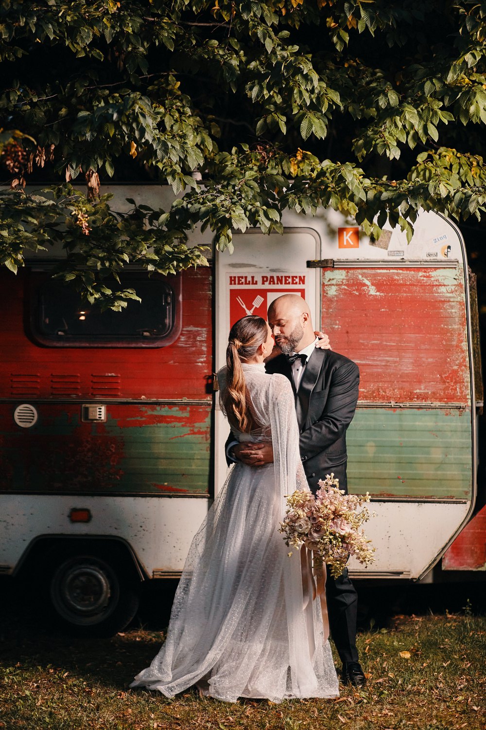 Gli sposi si baciano davanti a un vecchio camper, sotto un albero