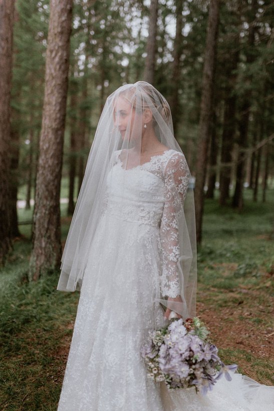 La sposa nel bosco di pini, il velo sul viso e un bouquet lilla